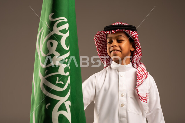 ذكرى العيد الوطني السعودي 23سبتمبر، الفخر والإعتزاز بإمتلاك الهوية الوطنية، حب الوطن والانتماء إليه، بورتريه مقرب لولد عربي خليجي سعودي يرتدي الثوب التقليدي والشماغ يحمل بيده علم المملكة وينظر إليه، الاحتفال بيوم العلم 11مارس، خلفية رمادية