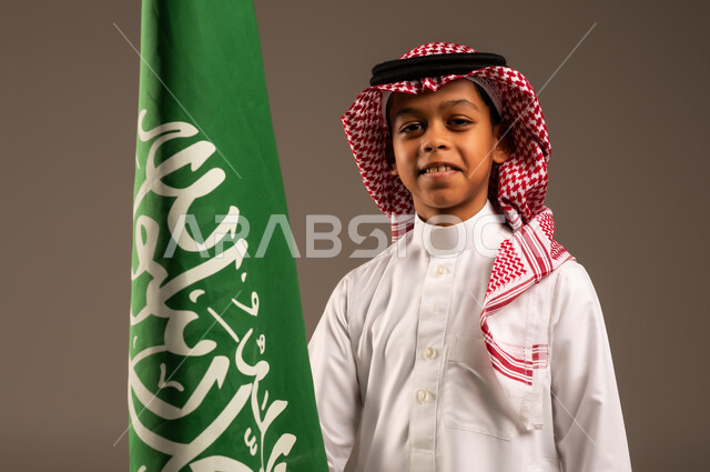 Saudi National Day 23 September, pride and honor in having a national identity, love of the homeland and belonging to it, close-up portrait of a Saudi Arabian Gulf boy wearing traditional dress and shemagh holding the Kingdom's flag in his hand and looking at the camera, celebrating Flag Day 11 March, gray background