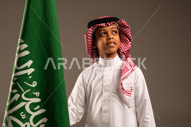 Saudi National Day 23 September, pride and honor in having a national identity, love of the homeland and belonging to it, close-up portrait of a Saudi Arabian Gulf boy wearing traditional dress and shemagh holding the Kingdom's flag in his hand and looking up, celebrating Flag Day 11 March, gray background