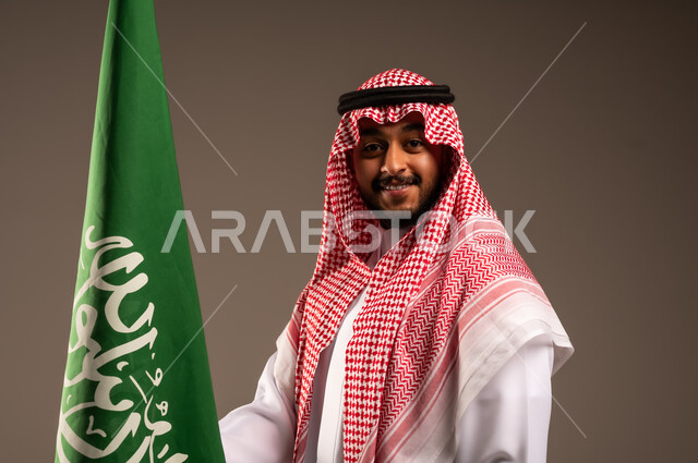 يوم العلم 11 مارس، الاحتفال بالعيد الوطني السعودي 23 سبتمبر، بورتريه مقرب لرجل عربي خليجي سعودي يرتدي الشماغ والثوب التقليدي يحمل بيده علم المملكة وينظر إلى الكاميرا، الفخر والاعتزاز بامتلاك الهوية الوطنية، خلفية رمادية