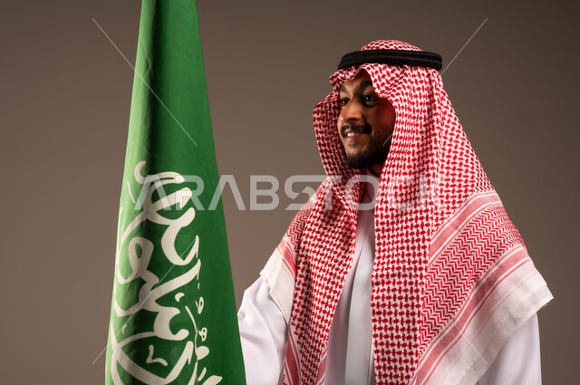 يوم العلم 11 مارس، الاحتفال بالعيد الوطني السعودي 23 سبتمبر، بورتريه مقرب لرجل عربي خليجي سعودي يرتدي الشماغ والثوب التقليدي يحمل بيده علم المملكة وينظر إليه، الفخر والاعتزاز بامتلاك الهوية الوطنية، خلفية رمادية