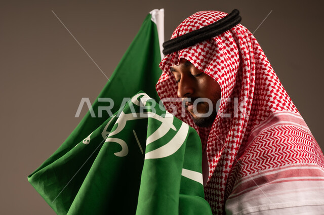 يوم العلم 11 مارس، الاحتفال بالعيد الوطني السعودي 23 سبتمبر، بورتريه مقرب لرجل عربي خليجي سعودي يرتدي الشماغ والثوب التقليدي يحمل بيده علم المملكة، الفخر والاعتزاز بامتلاك الهوية الوطنية، خلفية رمادية
