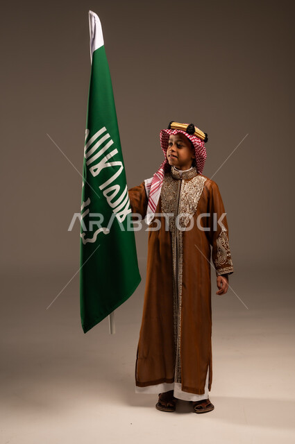 شعار الهوية الوطنية وراية الإسلام والتوحيد، إرتداء الأزياء الشعبية في الأعياد، الحب والإنتماء إلى الوطن، بورتريه لولد عربي خليجي سعودي يرتدي الدقلة والشماغ يحمل علم يوم التأسيس، يوم بدينا 1727 م، العيد الوطني السعودي 23سبتمبر، الطول الكامل للجسم، خلفية رمادية