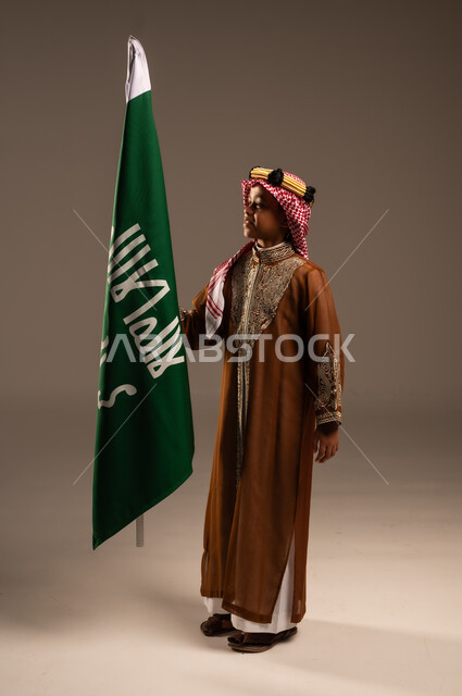 National identity logo and banner of Islam and monotheism, wearing traditional costumes on holidays, love and belonging to the homeland, portrait of a Saudi Gulf Arab boy wearing a dagla and a shemagh and kissing the flag of the founding day, the day we started 1727 AD, Saudi National Day September 23, full body length, gray background
