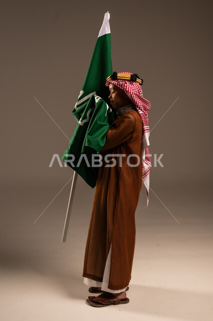 شعار الهوية الوطنية وراية الإسلام والتوحيد، إرتداء الأزياء الشعبية في الأعياد، الحب والإنتماء إلى الوطن، بورتريه لولد عربي خليجي سعودي يرتدي الدقلة والشماغ يقبل علم يوم التأسيس، يوم بدينا 1727 م، العيد الوطني السعودي 23سبتمبر، الطول الكامل للجسم، خلفية رمادية