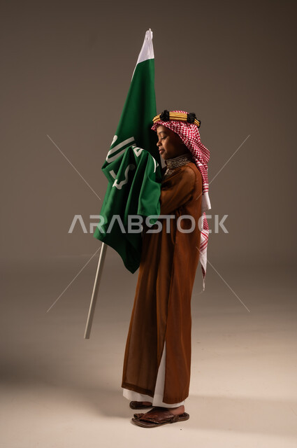 National identity logo and banner of Islam and monotheism, wearing traditional costumes on holidays, love and belonging to the homeland, portrait of a Saudi Gulf Arab boy wearing a dagla and a shemagh kissing the Founding Day flag, the day we started 1727 AD, Saudi National Day September 23, full body length, gray background