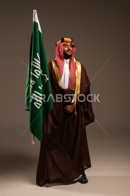 يوم بدينا 1727 م، شعار الهوية الوطنية وراية الإسلام والتوحيد، إرتداء الأزياء الشعبية في الأعياد، الحب والإنتماء إلى الوطن، بورتريه لرجل عربي خليجي سعودي يرتدي البشت والعقال يحمل علم يوم التأسيس، العيد الوطني السعودي 23سبتمبر، الطول الكامل للجسم، خلفية رمادية