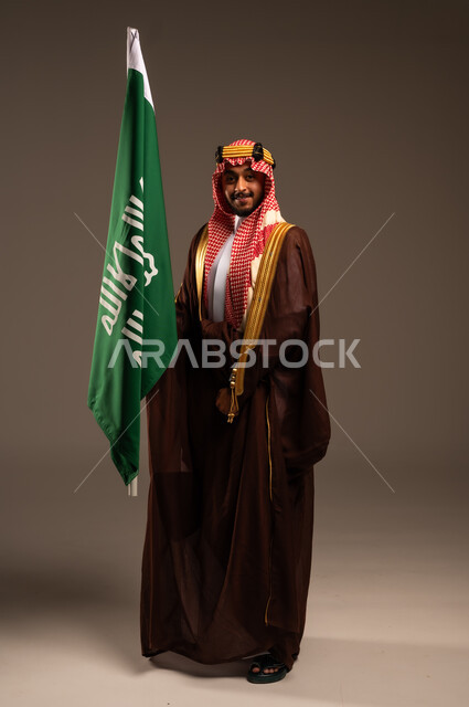 راية الإسلام والتوحيد وشعار الهوية الوطنية، يوم بدينا 1727 م، إرتداء الأزياء الشعبية في الأعياد، الحب والإنتماء إلى الوطن، بورتريه لرجل عربي خليجي سعودي يرتدي البشت والعقال يحمل علم يوم التأسيس، العيد الوطني السعودي 23سبتمبر، الطول الكامل للجسم، خلفية رمادية
