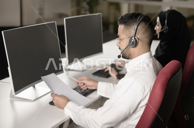 موظف عربي خليجي سعودي يعمل في خدمة العملاء ويمسك الاوراق بيده، موظف وموظفة يرتديان سماعة الاتصال، استعلامات واستفسارات، الاجابه على اسئلة العملاء، اجراء اتصال فيديو من خلال الحاسوب، متابعة وتطوير العمل