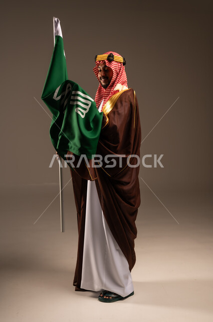 راية الإسلام والتوحيد وشعار الهوية الوطنية، يوم بدينا 1727 م، إرتداء الأزياء الشعبية في الأعياد، الحب والإنتماء إلى الوطن، بورتريه لرجل عربي خليجي سعودي يرتدي البشت والعقال يحمل علم يوم التأسيس، العيد الوطني السعودي 23سبتمبر، الطول الكامل للجسم، خلفية رمادية