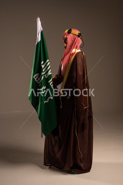 راية الإسلام والتوحيد وشعار الهوية الوطنية، يوم بدينا 1727 م، إرتداء الأزياء الشعبية في الأعياد، الحب والإنتماء إلى الوطن، بورتريه لرجل عربي خليجي سعودي يرتدي البشت والعقال يحمل علم يوم التأسيس، العيد الوطني السعودي 23سبتمبر، الطول الكامل للجسم، خلفية رمادية