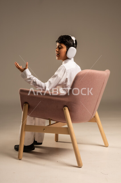 Using modern technical devices, listening to songs and music through headphones, portrait from the back of a Saudi Arabian Gulf child wearing headphones listening to something, full body, beige background