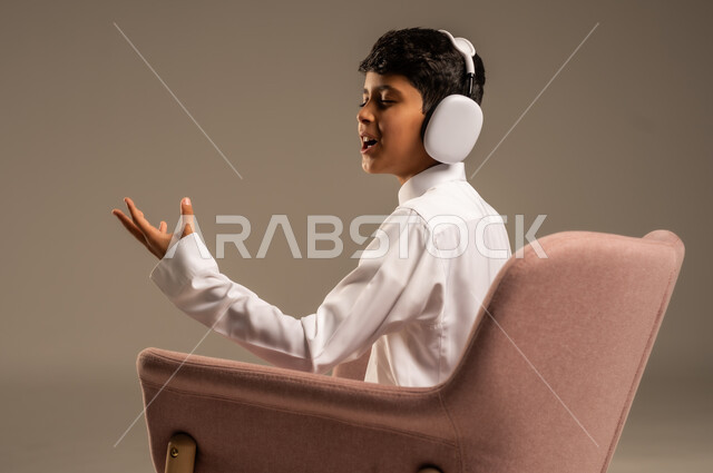 استخدام الأجهزة التقنية الحديثة، الاستماع الى الأغاني والموسيقى عبر سماعة الرأس، بورتريه من الخلف لطفل عربي خليجي سعودي يضع الهيدفون يقوم بالإصغاء إلى شيء ما، خلفية بيج