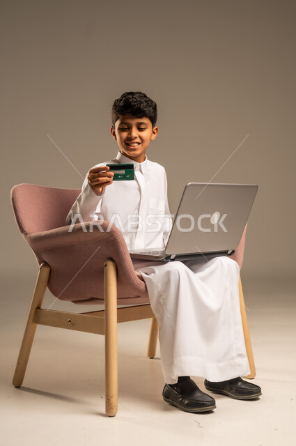 الشراء والدفع من خلال تطبيقات الجوال، التقنيات الحديثة والمتطورة، بورتريه لطفل عربي خليجي سعودي  يرتدي الثوب التقليدي يجلس على كرسي يمسك بيده الهاتف المحمول وبطاقة الإئتمان البنكية، تسديد الفواتير بالبطاقات الإلكترونية، الجسم بالطول الكامل، خلفية بيج