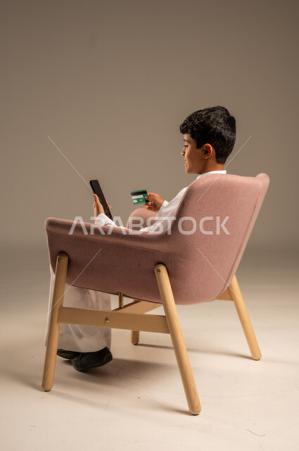 الشراء والدفع من خلال تطبيقات الجوال، التقنيات الحديثة والمتطورة، بورتريه لطفل عربي خليجي سعودي  يرتدي الثوب التقليدي يجلس على كرسي يمسك بيده الهاتف المحمول وبطاقة الإئتمان البنكية، تسديد الفواتير بالبطاقات الإلكترونية، الجسم بالطول الكامل، خلفية بيج