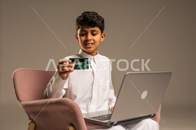 الشراء والدفع من خلال تطبيقات الجوال، التقنيات الحديثة والمتطورة، بورتريه مقرب لطفل عربي خليجي سعودي  يرتدي الثوب التقليدي يجلس على كرسي يمسك بيده الهاتف المحمول وبطاقة الإئتمان البنكية، تسديد الفواتير بالبطاقات الإلكترونية، خلفية بيج