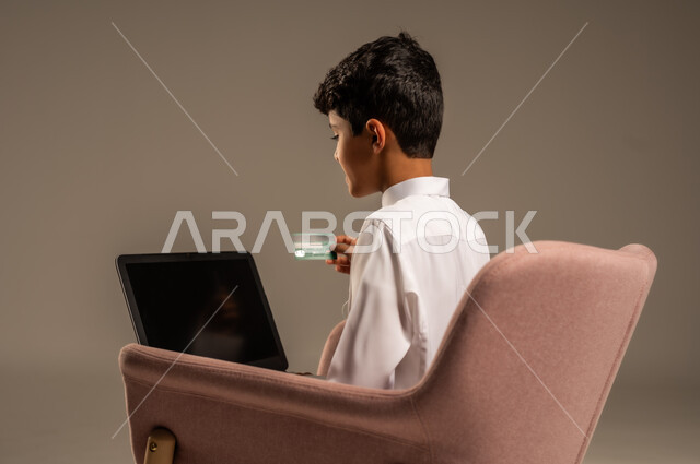 الشراء والدفع من خلال تطبيقات الجوال، التقنيات الحديثة والمتطورة، بورتريه مقرب لطفل عربي خليجي سعودي  يرتدي الثوب التقليدي يجلس على كرسي يمسك بيده الهاتف المحمول وبطاقة الإئتمان البنكية، تسديد الفواتير بالبطاقات الإلكترونية، خلفية بيج