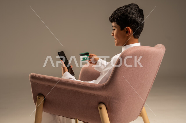 الشراء والدفع من خلال تطبيقات الجوال، التقنيات الحديثة والمتطورة، بورتريه مقرب لطفل عربي خليجي سعودي  يرتدي الثوب التقليدي يجلس على كرسي يمسك بيده الهاتف المحمول وبطاقة الإئتمان البنكية، تسديد الفواتير بالبطاقات الإلكترونية، خلفية بيج