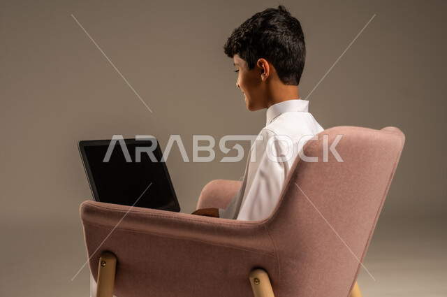 محادثات ودردشة عبر الانترتت, بورتريه من الخلف لطفل عربي سعودي خليجي يرتدي الثوب السعودي ويجلس على كرسي مريح يقوم بإستخدام جهاز الحاسوب المحمول، التواصل مع الأصدقاء عن بعد، مفهوم التعلم اونلاين، قضاء أوقات ممتعة على اللابتوب، خلفية بيج