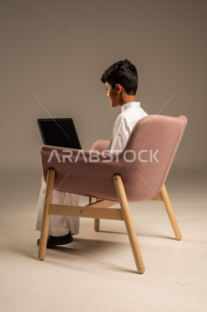 محادثات ودردشة عبر الانترتت, بورتريه من الخلف لطفل عربي سعودي خليجي يرتدي الثوب السعودي ويجلس على كرسي مريح يقوم بإستخدام جهاز الحاسوب المحمول، التواصل مع الأصدقاء عن بعد، مفهوم التعلم اونلاين، قضاء أوقات ممتعة على اللابتوب، خلفية بيج
