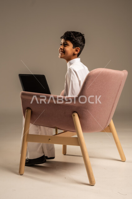 محادثات ودردشة عبر الانترتت, بورتريه من الخلف لطفل عربي سعودي خليجي يرتدي الثوب السعودي ويجلس على كرسي مريح يقوم بإستخدام جهاز الحاسوب المحمول، التواصل مع الأصدقاء عن بعد، مفهوم التعلم اونلاين، قضاء أوقات ممتعة على اللابتوب، خلفية بيج