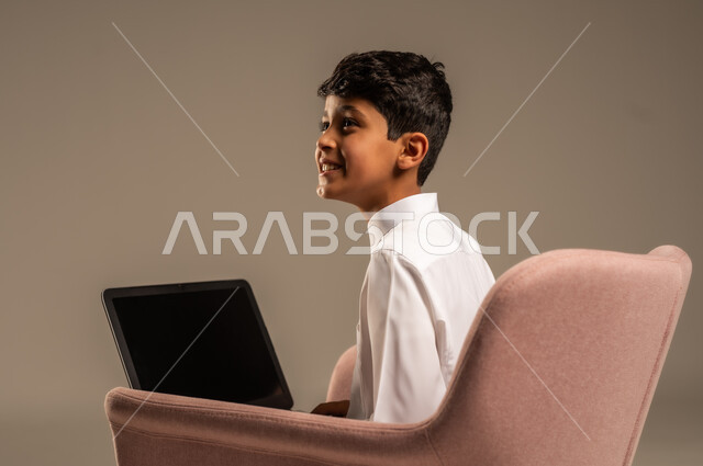 محادثات ودردشة عبر الانترتت, بورتريه من الخلف لطفل عربي سعودي خليجي يرتدي الثوب السعودي ويجلس على كرسي مريح يقوم بإستخدام جهاز الحاسوب المحمول، التواصل مع الأصدقاء عن بعد، مفهوم التعلم اونلاين، قضاء أوقات ممتعة على اللابتوب، خلفية بيج