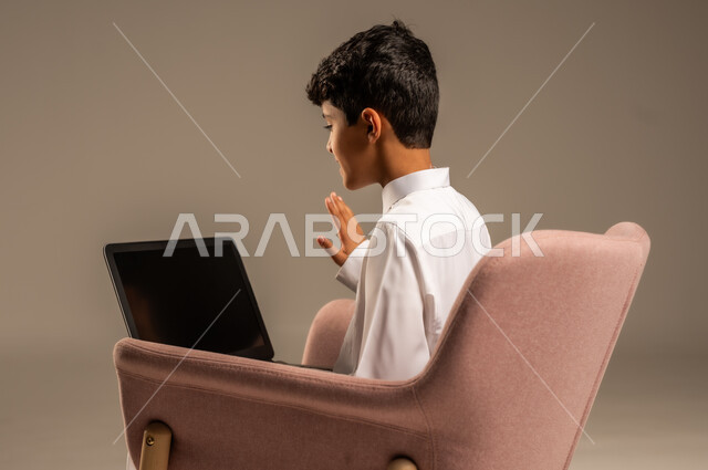 محادثات ودردشة عبر الانترتت, بورتريه من الخلف لطفل عربي سعودي خليجي يرتدي الثوب السعودي ويجلس على كرسي مريح يقوم بإستخدام جهاز الحاسوب المحمول، التواصل مع الأصدقاء عن بعد، مفهوم التعلم اونلاين، قضاء أوقات ممتعة على اللابتوب، خلفية بيج
