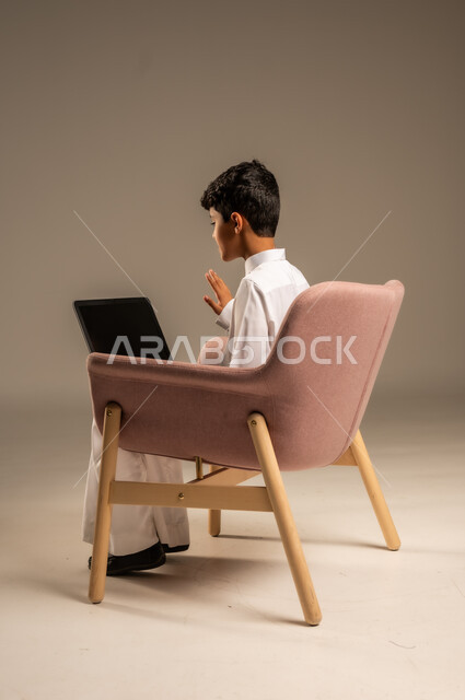 محادثات ودردشة عبر الانترتت, بورتريه من الخلف لطفل عربي سعودي خليجي يرتدي الثوب السعودي ويجلس على كرسي مريح يقوم بإستخدام جهاز الحاسوب المحمول، التواصل مع الأصدقاء عن بعد، مفهوم التعلم اونلاين، قضاء أوقات ممتعة على اللابتوب، خلفية بيج