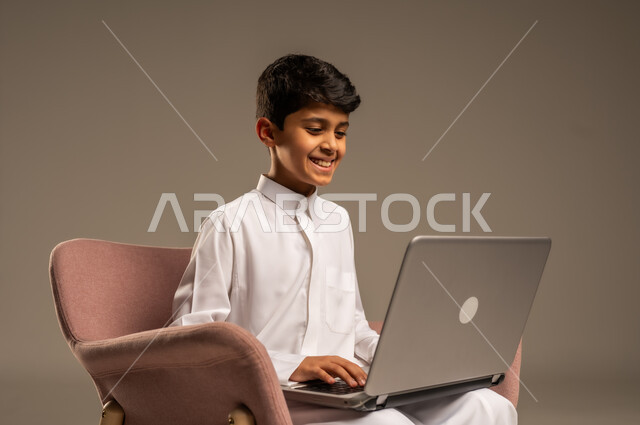 التواصل مع الأصدقاء عن بعد، محادثات ودردشة عبر الانترتت, بورتريه  لطفل عربي سعودي خليجي يرتدي الثوب السعودي ويجلس على كرسي مريح يقوم بإستخدام جهاز الحاسوب المحمول، مفهوم التعلم اونلاين، قضاء أوقات ممتعة على اللابتوب، خلفية بيج