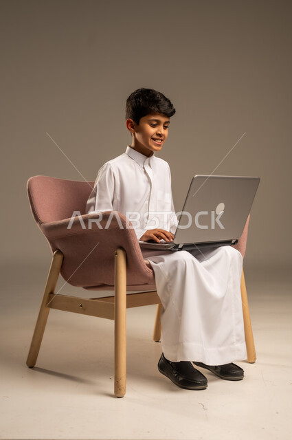 التواصل مع الأصدقاء عن بعد، محادثات ودردشة عبر الانترتت, بورتريه  لطفل عربي سعودي خليجي يرتدي الثوب السعودي ويجلس على كرسي مريح يقوم بإستخدام جهاز الحاسوب المحمول، مفهوم التعلم اونلاين، قضاء أوقات ممتعة على اللابتوب، خلفية بيج