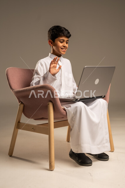 Communicating with friends remotely, online conversations and chat, portrait of a Saudi Arabian Gulf child wearing a Saudi thobe and sitting on a comfortable chair using a laptop, online learning concept, having fun on the laptop, beige background