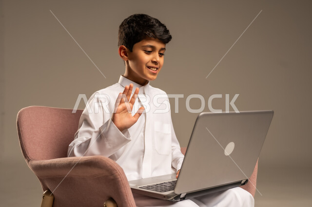التواصل مع الأصدقاء عن بعد، محادثات ودردشة عبر الانترتت, بورتريه  لطفل عربي سعودي خليجي يرتدي الثوب السعودي ويجلس على كرسي مريح يقوم بإستخدام جهاز الحاسوب المحمول، مفهوم التعلم اونلاين، قضاء أوقات ممتعة على اللابتوب، خلفية بيج