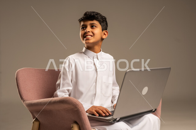 التواصل مع الأصدقاء عن بعد، محادثات ودردشة عبر الانترتت, بورتريه  لطفل عربي سعودي خليجي يرتدي الثوب السعودي ويجلس على كرسي مريح يقوم بإستخدام جهاز الحاسوب المحمول، مفهوم التعلم اونلاين، قضاء أوقات ممتعة على اللابتوب، خلفية بيج