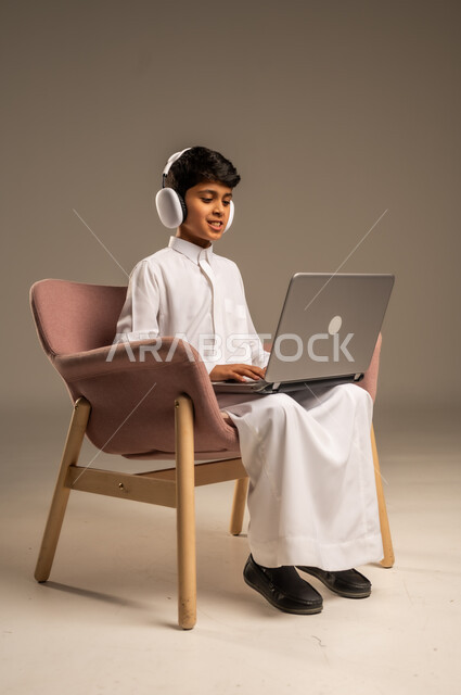 قضاء أوقات ممتعة على اللابتوب،  التواصل مع الأصدقاء عن بعد، محادثات ودردشة عبر الانترتت, بورتريه  لطفل عربي سعودي خليجي يرتدي الثوب السعودي ويجلس على كرسي مريح يقوم بإستخدام جهاز الحاسوب المحمول ويضع سماعات الرأس، مفهوم التعلم اونلاين، خلفية بيج