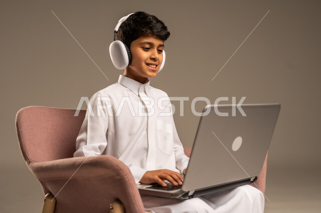  قضاء أوقات ممتعة على اللابتوب،  التواصل مع الأصدقاء عن بعد، محادثات ودردشة عبر الانترتت, بورتريه  لطفل عربي سعودي خليجي يرتدي الثوب السعودي ويجلس على كرسي مريح يقوم بإستخدام جهاز الحاسوب المحمول ويضع سماعات الرأس، مفهوم التعلم اونلاين، خلفية بيج