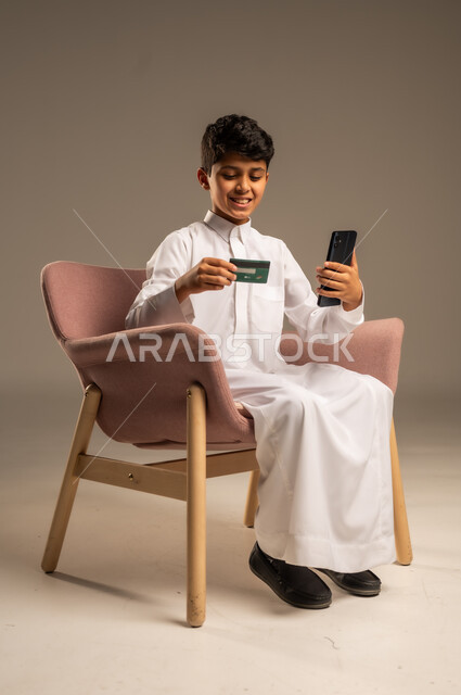 مفهوم الدفع والتسديد عن بعد عبر تطبيقات الجوال، التسوق الالكتروني عبر الانترنت، استخدام البطاقة البنكية الالكترونية، بورتريه لطفل عربي سعودي خليجي  يرتدي الثوب التقليدي  يجلس على كرسي مريح يحمل الهاتف المحمول، خلفية بيج