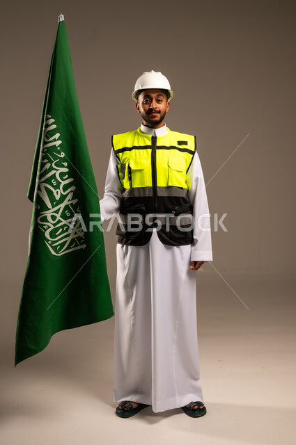 Participating in the celebration of national occasions and holidays, commemorating the Saudi National Day, September 23, portrait of a Saudi Arabian Gulf engineer wearing traditional dress, jacket and protective hat holding the flag of the Kingdom of Saudi Arabia with gestures of pride and honor, full body, gray background