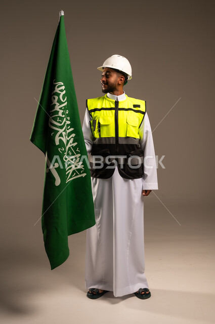 المشاركة بالاحتفال بالمناسبات والأعياد الوطنية، إحياء ذكرى اليوم الوطني السعودي 23سبتمبر، بورتريه لمهندس عربي خليجي سعودي يرتدي الثوب التقليدي والسترة وقبعة الحماية يحمل راية المملكة العربية السعودية بإيماءات الفخر والاعتزاز، الجسم بالطول الكامل، خلفية رمادية