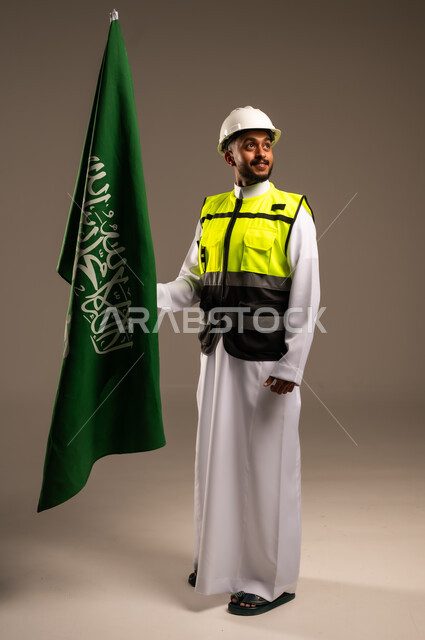Participating in the celebration of national occasions and holidays, commemorating the Saudi National Day, September 23, portrait of a Saudi Arabian Gulf engineer wearing traditional dress, jacket and protective hat holding the flag of the Kingdom of Saudi Arabia with gestures of pride and honor, full body, gray background