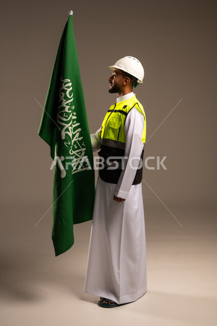 المشاركة بالاحتفال بالمناسبات والأعياد الوطنية، إحياء ذكرى اليوم الوطني السعودي 23سبتمبر، بورتريه لمهندس عربي خليجي سعودي يرتدي الثوب التقليدي والسترة وقبعة الحماية يحمل راية المملكة العربية السعودية بإيماءات الفخر والاعتزاز، الجسم بالطول الكامل، خلفية رمادية