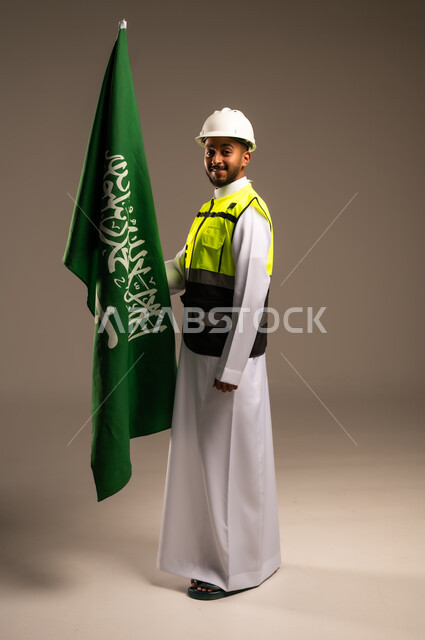 المشاركة بالاحتفال بالمناسبات والأعياد الوطنية، إحياء ذكرى اليوم الوطني السعودي 23سبتمبر، بورتريه لمهندس عربي خليجي سعودي يرتدي الثوب التقليدي والسترة وقبعة الحماية يحمل راية المملكة العربية السعودية بإيماءات الفخر والاعتزاز، الجسم بالطول الكامل، خلفية رمادية