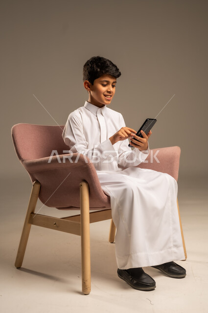 Using advanced modern technology and technical devices, gestures of happiness and joy, close-up portrait of a smiling Saudi Arabian Gulf child wearing traditional dress holding a mobile phone in his hand, beige background