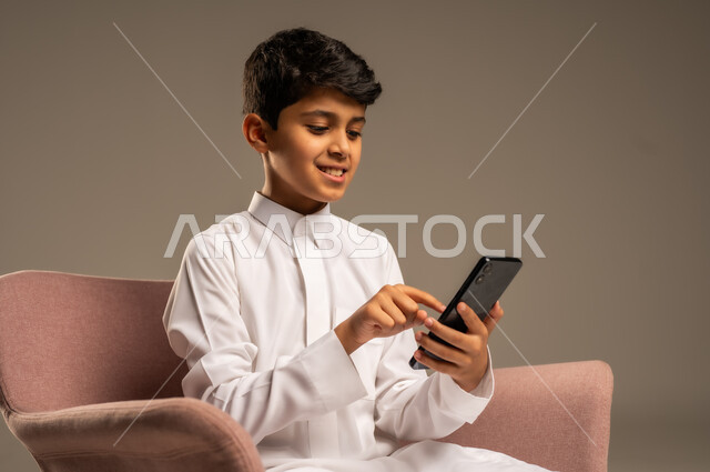 Using advanced modern technology and technical devices, gestures of happiness and joy, close-up portrait of a smiling Saudi Arabian Gulf child wearing traditional dress holding a mobile phone in his hand, beige background