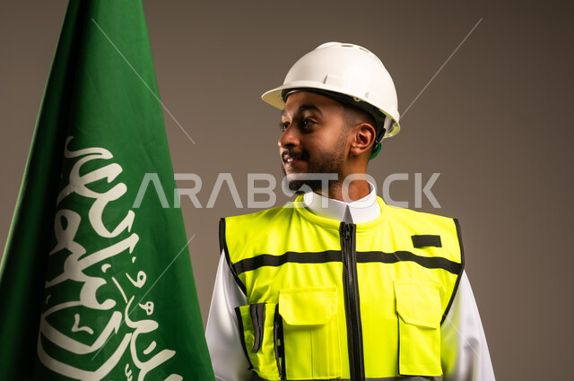 Celebrating Saudi National Day September 23, Participating in the celebration of national occasions and holidays, Close-up portrait of a Saudi Arabian Gulf engineer wearing traditional dress, jacket and protective hat holding the flag of the Kingdom of Saudi Arabia with gestures of pride and honor, gray background