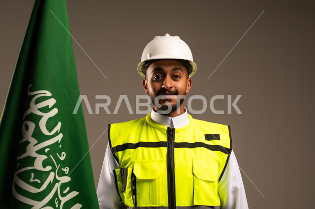 Celebrating Saudi National Day September 23, Participating in the celebration of national occasions and holidays, Close-up portrait of a Saudi Arabian Gulf engineer wearing traditional dress, jacket and protective hat holding the flag of the Kingdom of Saudi Arabia with gestures of pride and honor, gray background