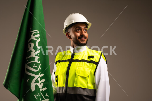 Celebrating Saudi National Day September 23, Participating in the celebration of national occasions and holidays, Close-up portrait of a Saudi Arabian Gulf engineer wearing traditional dress, jacket and protective hat holding the flag of the Kingdom of Saudi Arabia with gestures of pride and honor, gray background