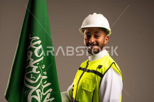 Celebrating Saudi National Day September 23, Participating in the celebration of national occasions and holidays, Close-up portrait of a Saudi Arabian Gulf engineer wearing traditional dress, jacket and protective hat holding the flag of the Kingdom of Saudi Arabia with gestures of pride and honor, gray background