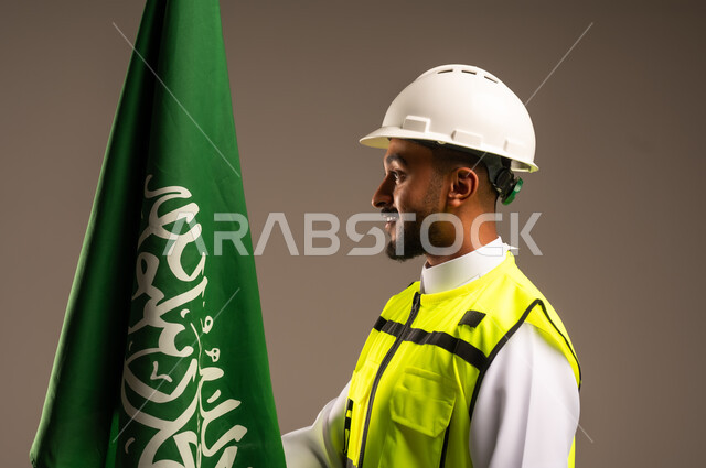 Celebrating Saudi National Day September 23, Participating in the celebration of national occasions and holidays, Close-up portrait of a Saudi Arabian Gulf engineer wearing traditional dress, jacket and protective hat holding the flag of the Kingdom of Saudi Arabia with gestures of pride and honor, gray background