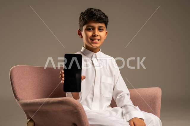استخدام التكنولوجيا والأجهزة التقنية الحديثة المتطورة، ايماءات السعادة والفرح، بورتريه مقرب لطفل عربي خليجي سعودي مبتسم يرتدي الثوب التقليدي يمسك بيده الهاتف المحمول، خلفية بيج