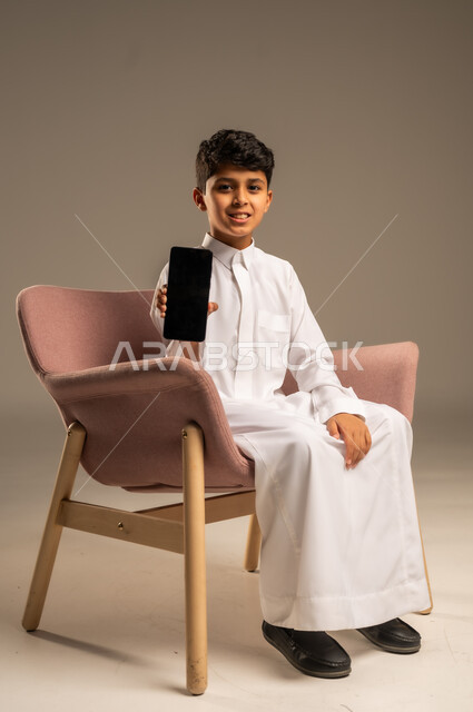 استخدام التكنولوجيا والأجهزة التقنية الحديثة المتطورة، ايماءات السعادة والفرح، بورتريه مقرب لطفل عربي خليجي سعودي مبتسم يرتدي الثوب التقليدي يمسك بيده الهاتف المحمول، خلفية بيج